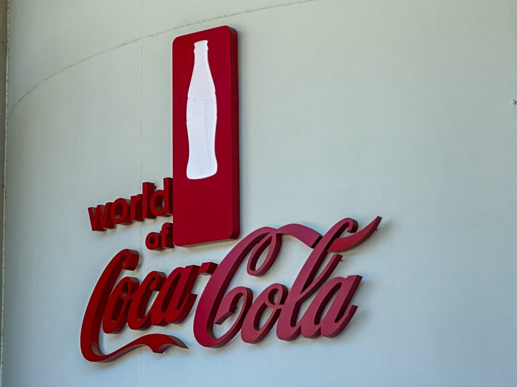 Alt text: A sign on a wall reads "World of Coca-Cola" with the words "World of" in smaller red letters and "Coca-Cola" in larger, stylized red script. Above the text is a red rectangular sign with a white silhouette of a Coca-Cola bottle.