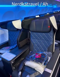 The image shows a row of business or first-class airplane seats. The seats are upholstered in dark blue leather with a diamond pattern and have headrests with white covers. Each seat has a large screen in front of it and a tray table. There are packaged blankets and headphones placed on the seats. The cabin lighting has a blue hue, giving the area a modern and comfortable ambiance.