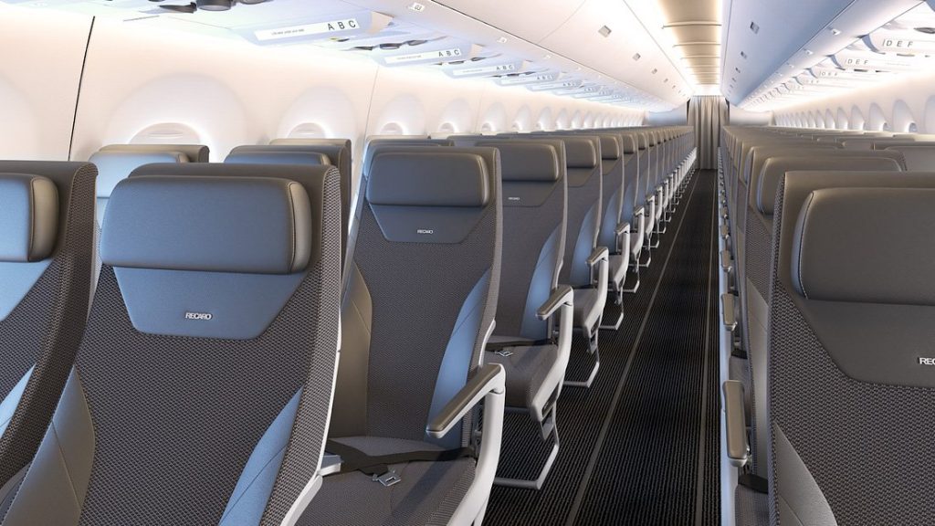 The image shows the interior of an airplane cabin. The cabin features rows of modern, grey seats with headrests, arranged in a 3-3 configuration on either side of a central aisle. The overhead compartments are closed, and the lighting is bright, giving the cabin a clean and spacious appearance. The seats are labeled with the brand "RECARO."