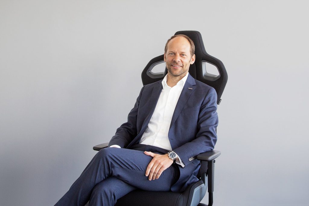 A man in a blue suit and white dress shirt is sitting in a black office chair against a plain gray background. He is smiling and has his legs crossed, with his hands resting on his lap.