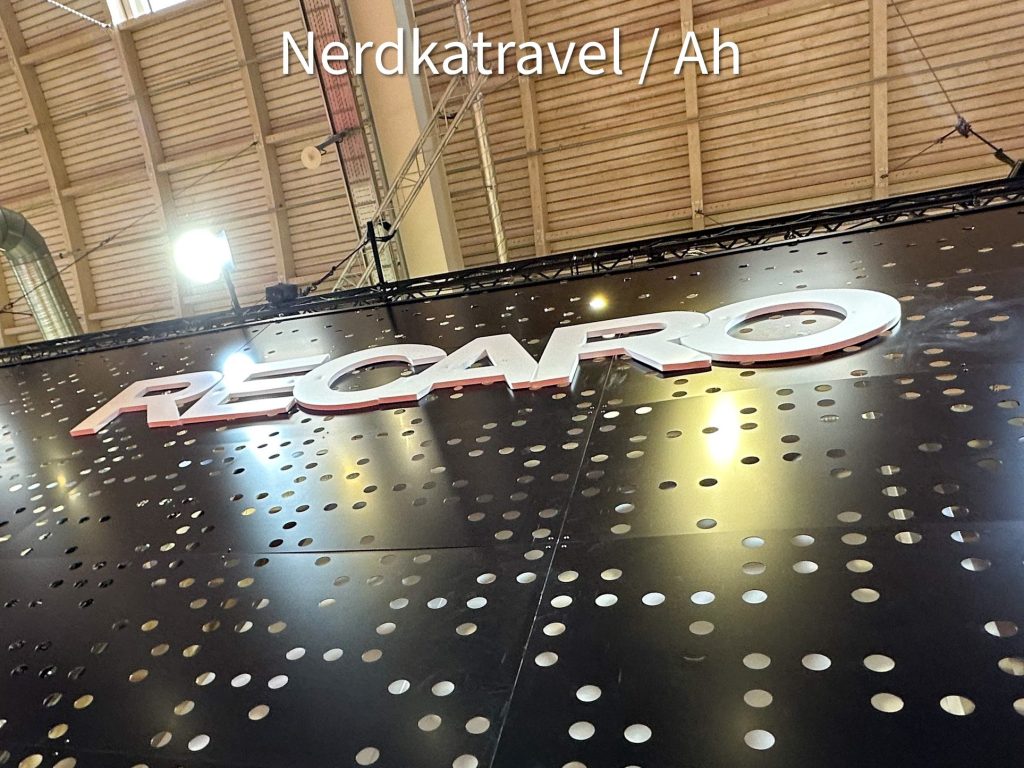 Alt text: A large sign with the word "RECARO" in bold white letters is displayed on a black perforated background. The image is taken from a low angle, showing the ceiling of the building with lights and structural elements. The text "Nerdkatravel / Ah" is overlaid at the top of the image.