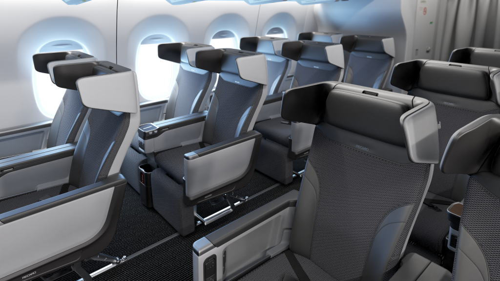 The image shows the interior of an airplane cabin, specifically the premium economy or business class section. The seats are arranged in a 2-2 configuration, with each seat featuring a large headrest, ample legroom, and individual armrests. The seats are upholstered in a combination of dark and light gray fabric, with some metallic accents. The cabin has large windows, and the overall design appears modern and comfortable.