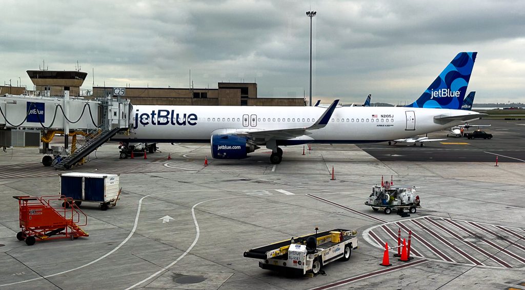 A JetBlue airplane is parked at an airport gate, connected to a jet bridge labeled "C25." The aircraft is white with blue accents and has "jetBlue" written on the side and tail. Various ground service equipment, including a baggage cart, a pushback tug, and other utility vehicles, are positioned around the plane. The sky is overcast, and the airport terminal buildings are visible in the background.