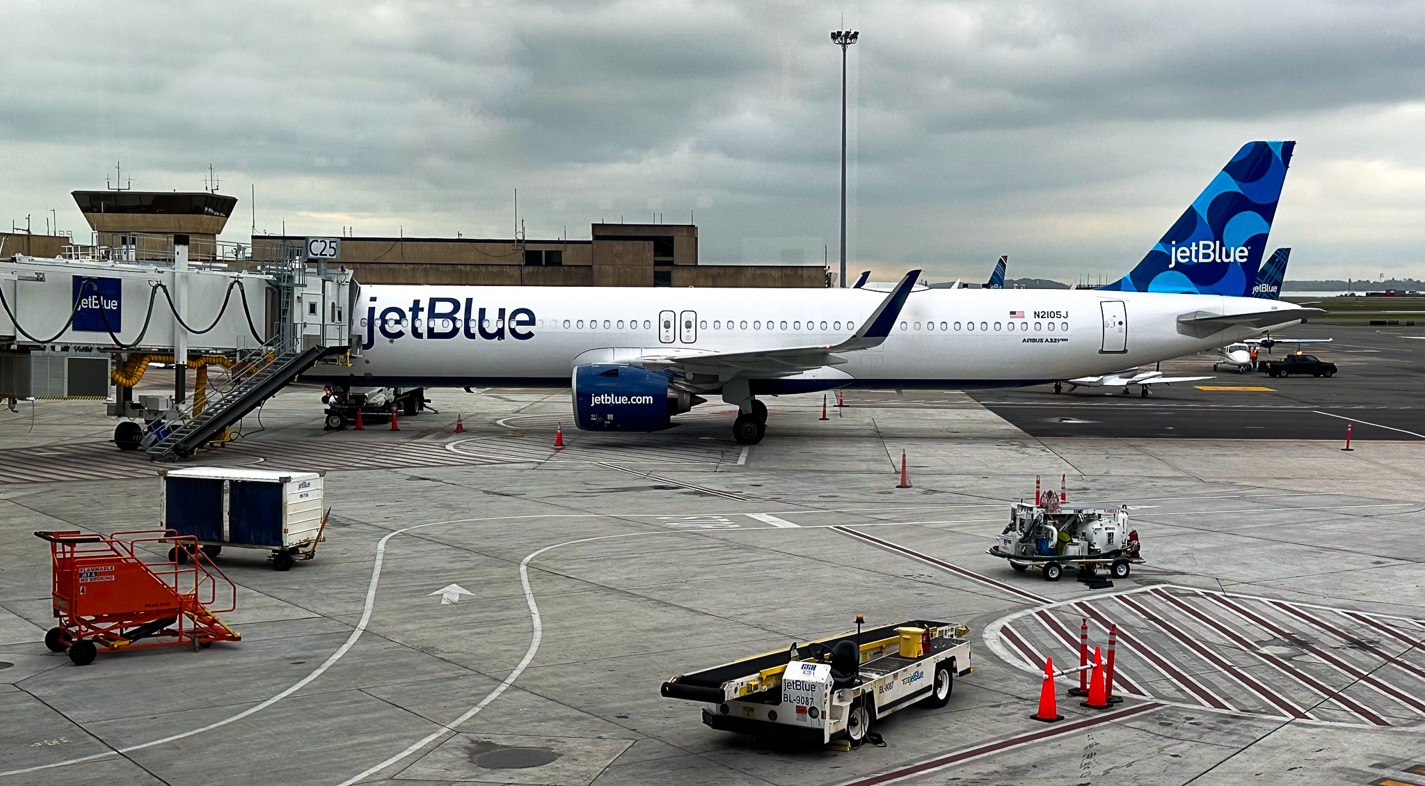 A JetBlue airplane is parked at an airport gate, connected to a jet bridge labeled "C25." The aircraft is white with blue accents and has "jetBlue" written on the side and tail. Various ground service equipment, including a baggage cart, a pushback tug, and other utility vehicles, are positioned around the plane. The sky is overcast, and the airport terminal buildings are visible in the background.