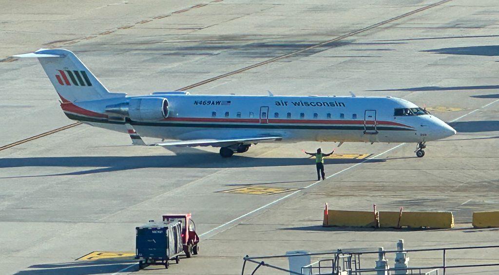 The image shows an Air Wisconsin airplane on the tarmac at an airport. The aircraft is a regional jet with the registration number N469AW. A ground crew member in a high-visibility vest is standing in front of the plane, directing it with hand signals. There is ground support equipment, including a baggage cart, visible near the plane. The scene is set in daylight.