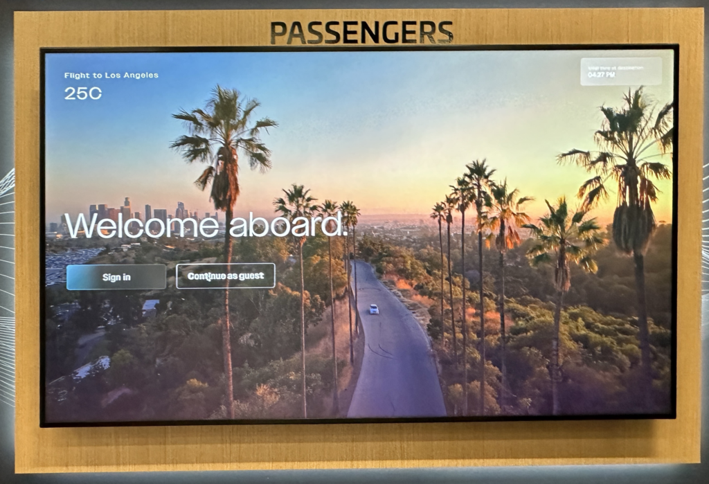 The image shows a digital screen with a wooden frame labeled "PASSENGERS" at the top. The screen displays a scenic view of a road lined with tall palm trees, leading towards a city skyline in the distance, likely Los Angeles, as indicated by the text. The sky is clear with a warm, golden hue, suggesting either sunrise or sunset. The screen has text that reads "Flight to Los Angeles," "25C," "Welcome aboard," and options to "Sign in" or "Continue as guest." The top right corner shows the local time at the destination as 04:27 PM.
