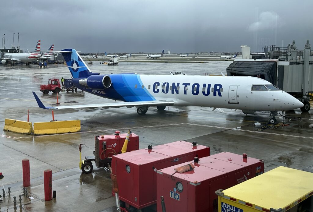 The image shows a Contour Airlines jet parked at an airport gate. The aircraft is white with blue accents and the word "CONTOUR" written in large blue letters on the side. The tail of the plane features a blue design with a circular logo. The scene is wet, indicating recent rain, and there are various ground service equipment and personnel around the plane. In the background, other aircraft from different airlines are visible on the tarmac. The sky is overcast, contributing to the overall gray and rainy atmosphere.