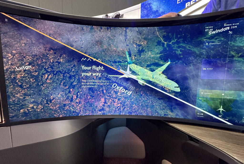 A curved screen displays a flight map showing the current position of an airplane over a geographical area. The map highlights locations such as Luton and Oxford. The screen also provides flight information, including the current time in Los Angeles (07:30), the estimated arrival time in Los Angeles (20 minutes), and the flight path. The text on the screen reads "Your flight, your way" and suggests exploring the in-flight services available. The map also indicates meal times and sleep periods during the flight. The screen is part of an airplane's in-flight entertainment system, and part of a seat and table are visible in the foreground.