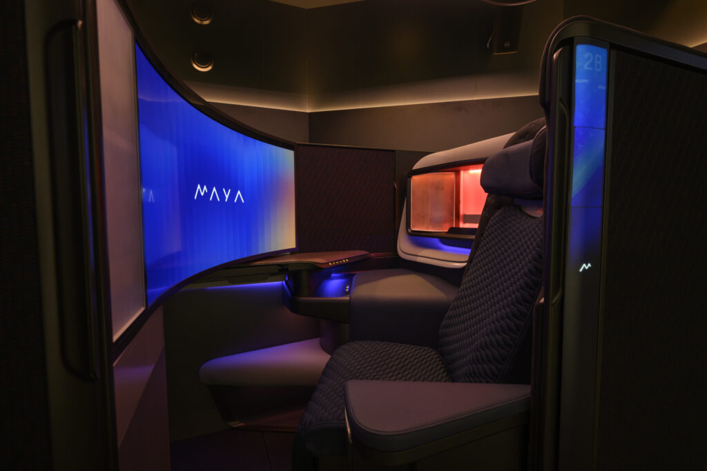 The image shows a luxurious, private seating area, likely in a first-class airplane cabin or a high-end lounge. The space features a large, curved screen displaying the word "MAYA" in white text on a blue background. The seat is padded and upholstered in a dark, textured fabric, with a headrest and armrests. The area is dimly lit with ambient lighting, including a red light source behind a partition. The overall design is modern and sleek, emphasizing comfort and privacy.