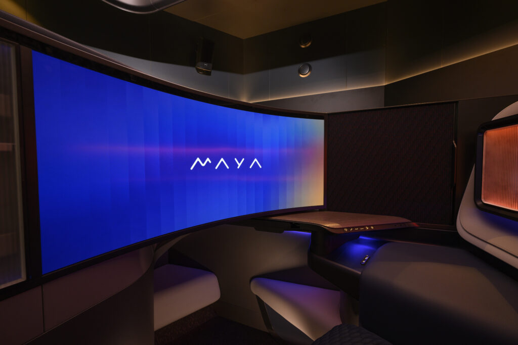 The image shows a modern, high-tech workspace or entertainment setup with a large, curved screen displaying the word "MAYA" in the center. The screen is illuminated with a gradient of blue and purple hues. The surrounding area includes a sleek, ergonomic desk and seating, with ambient lighting that adds to the futuristic and sophisticated atmosphere.