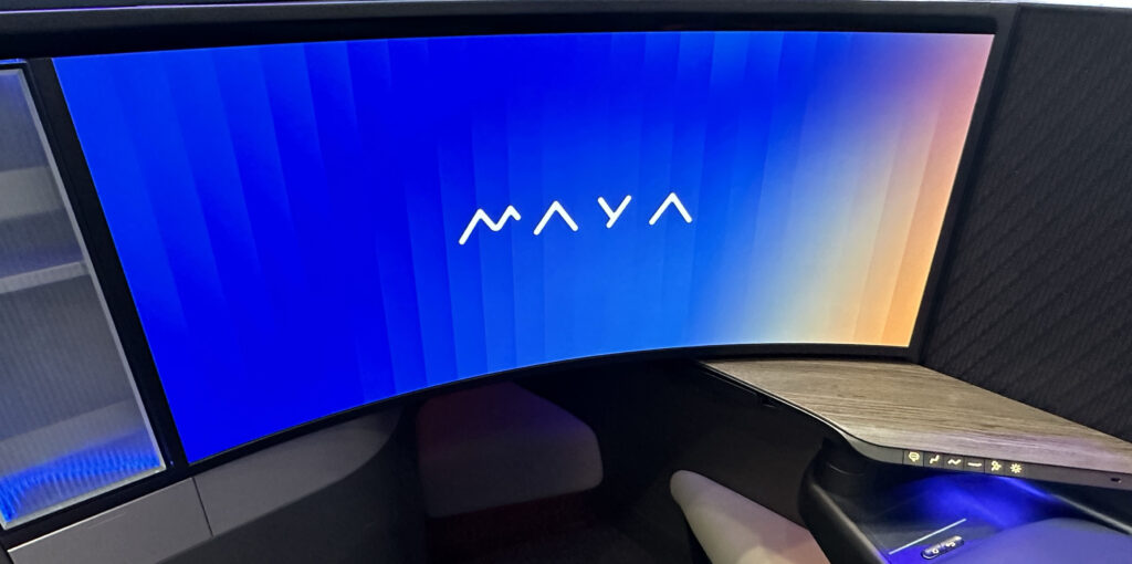 The image shows a modern, curved computer monitor displaying the word "MAYA" in white text against a gradient background that transitions from blue on the left to orange on the right. The monitor is part of a sleek, high-tech workspace setup, which includes a wooden desk surface and a control panel with various icons and buttons.